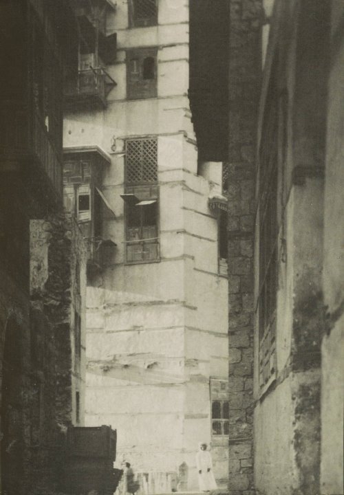 Photograph of a person in all white Arabian garment in the distance, facing the camera, with tall buildings beside and behind them.