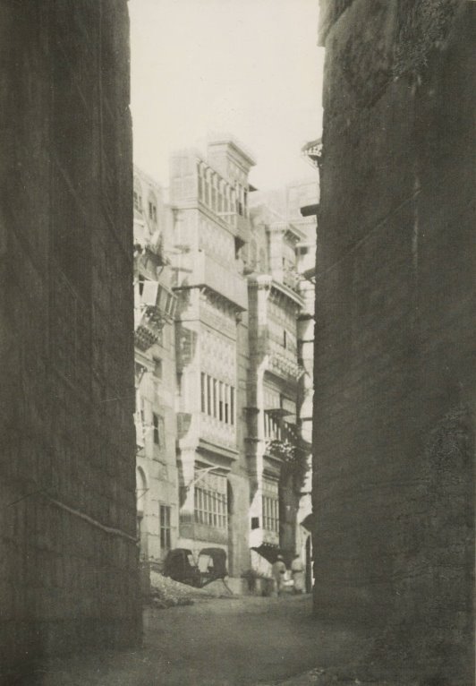 Photograph of an alley with two tall buildings on either side, opening into an area with several tall light-coloured buildings.