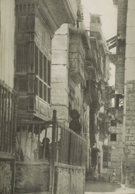 Photograph of a narrow street, with two people sitting on an enclosed balcony on the left, and a person facing the camera off in the distance.