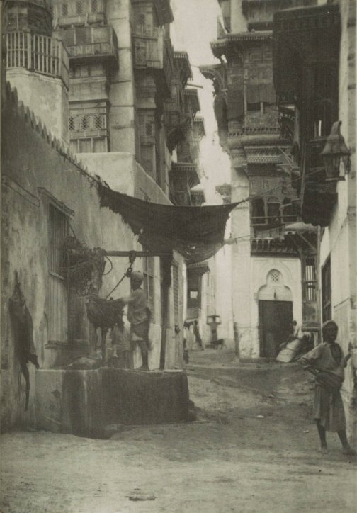 Photograph of a narrow street, with a woman on the right standing next to a building, and a person on the left on a raised area, looking at the camera.