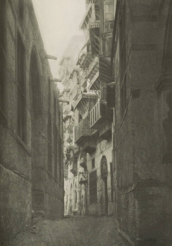 Photograph of a very narrow street with tall dark buildings on either side and lighter buildings beyond as the street curves to the left.