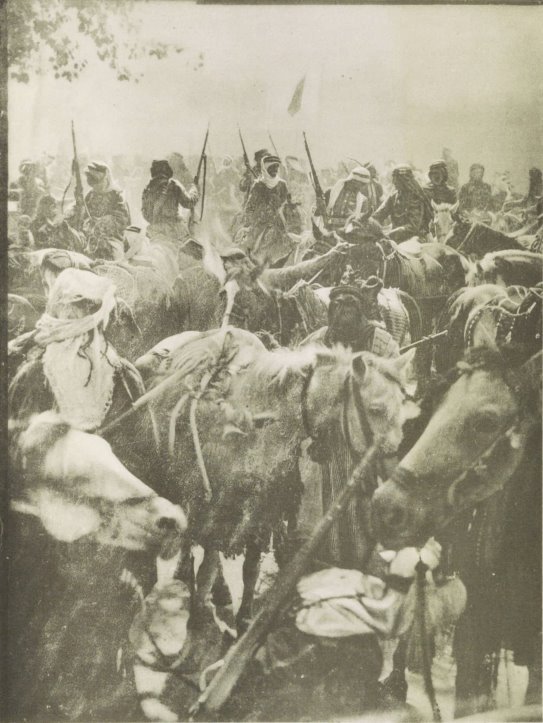 Photograph of Lawrence and his men on horses, with others walking, as they come into the city of Damascus.